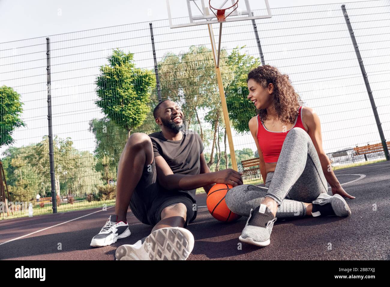 Attività all'aperto. Coppia africana seduta con palla sul campo di pallacanestro che parla sorridente allegro Foto Stock