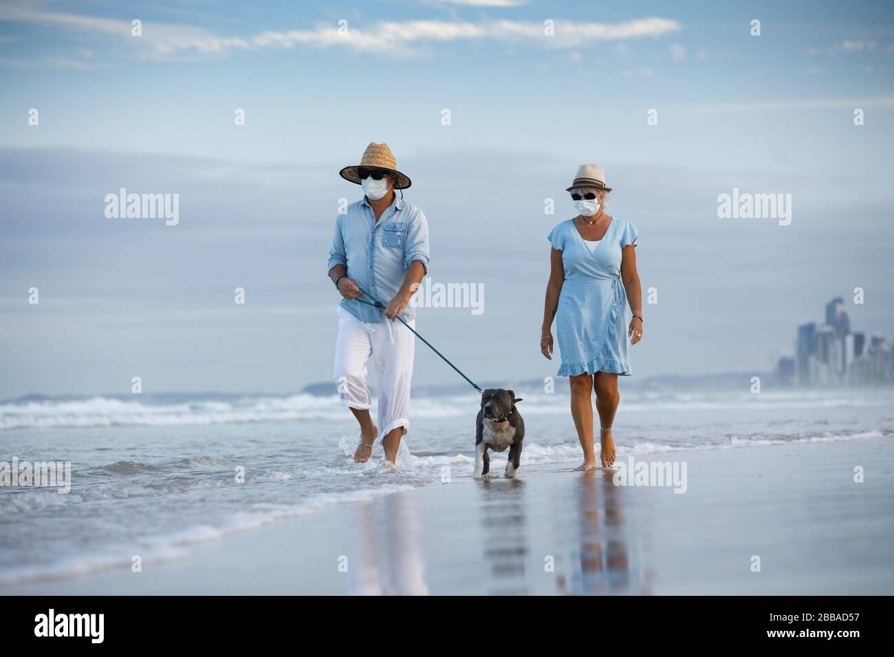 una coppia con maschere mediche che camminano un mare blu cane stafford su una spiaggia bella. concetto di cane a piedi o rimanere attivo in mezzo a coronavirus pandemic Foto Stock