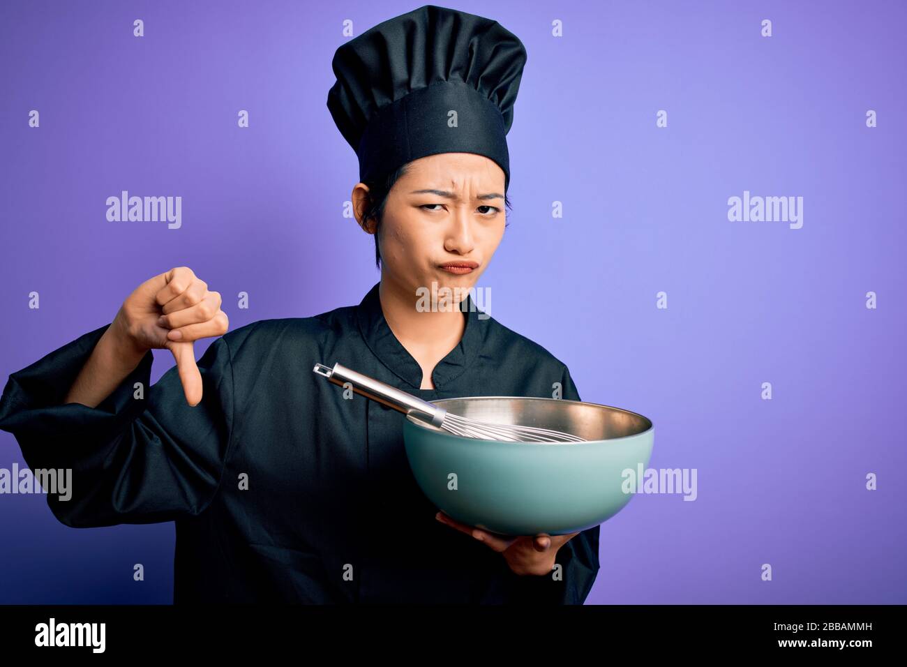 Giovane e bella donna chef cinese che indossa uniforme cucina e cappello  utilizzando ciotola e sussurra con viso arrabbiato, segno negativo  mostrando dispiace con pollici giù Foto stock - Alamy