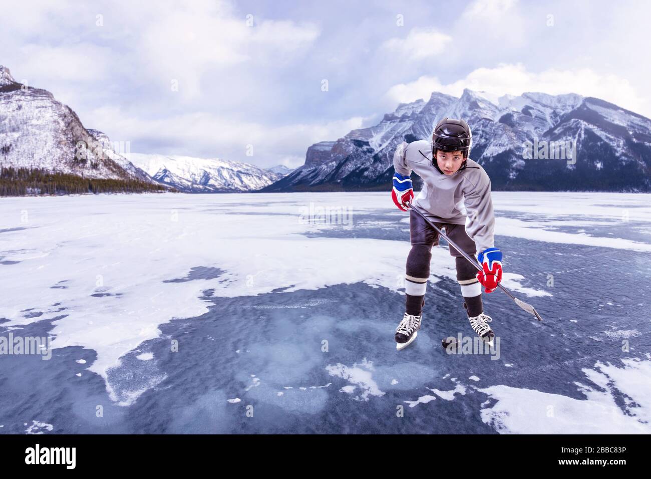 Giocatore di hockey su ghiaccio Junior indossando casco, maglia e tenendo il bastone da hockey su disco in azione all'aperto circondato dalle montagne delle Montagne Rocciose canadesi. Foto Stock