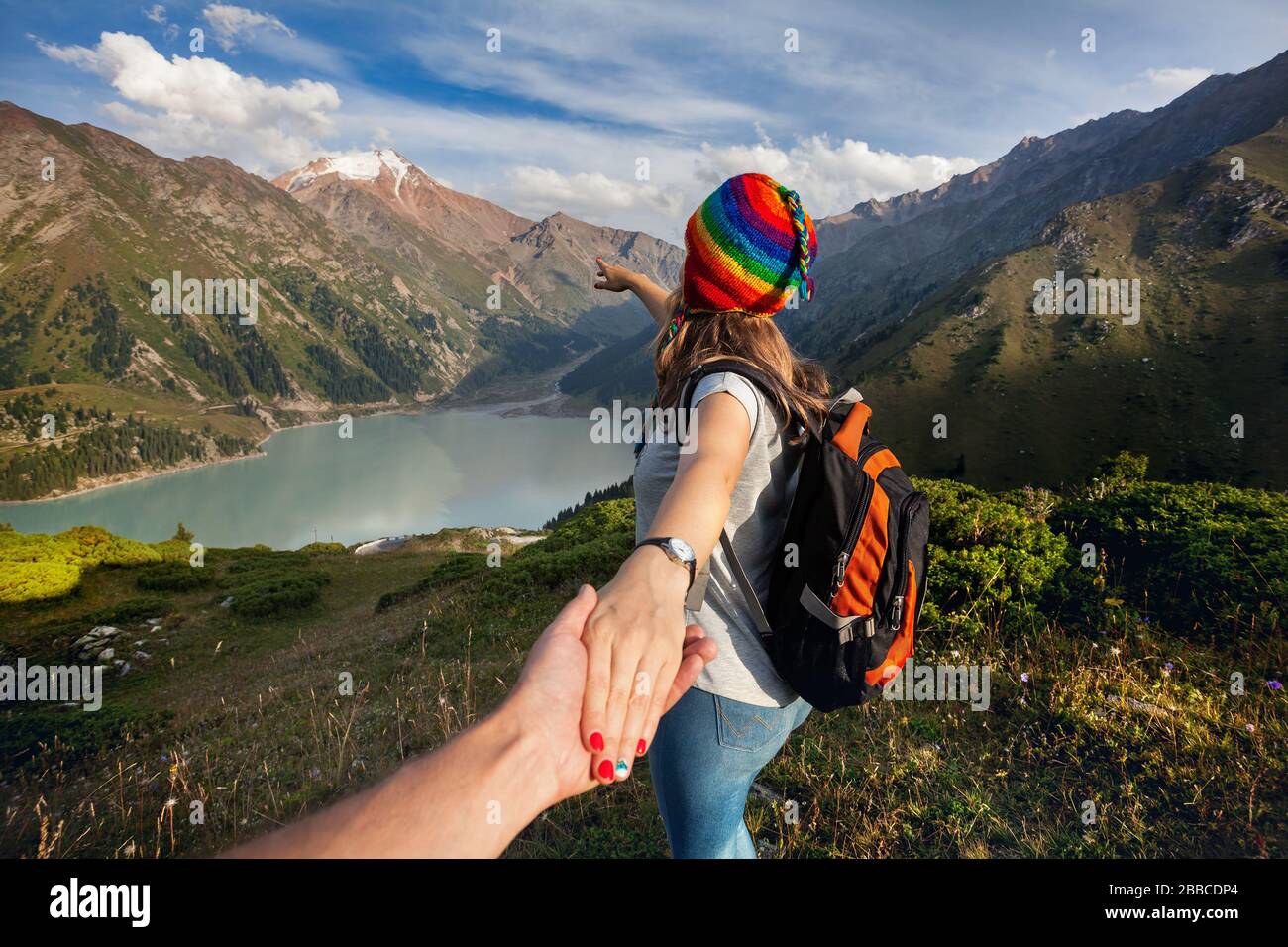 Donna turistica in arcobaleno hat e poncho marrone azienda man a mano e rivolta verso il lago di montagna Foto Stock