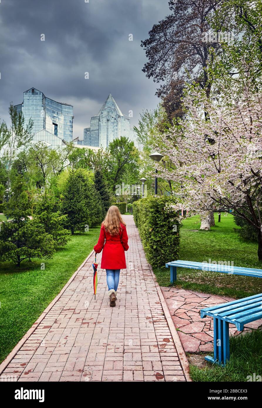 Woman in Red cappotto e ombrello a piedi giù per la strada nel parco con fiori di ciliegio e cielo nuvoloso Foto Stock