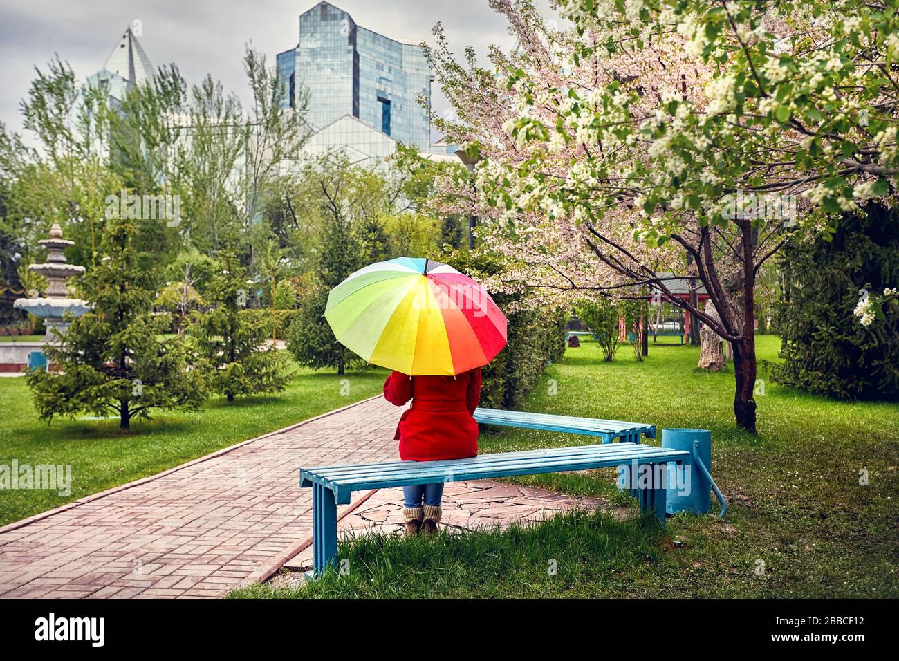 Woman in Red coat con ombrello arcobaleno seduta nel parco con alberi in fiore Foto Stock