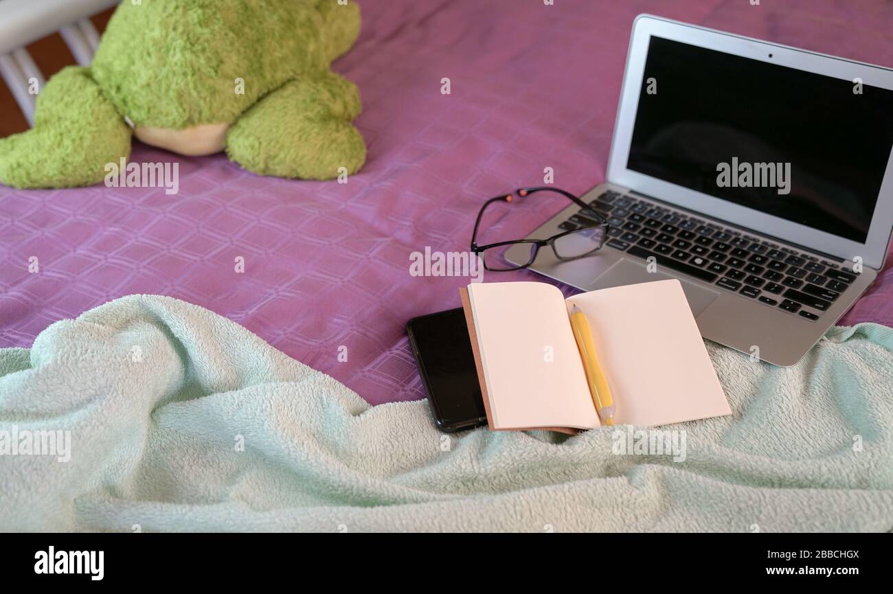 Computer portatile con blocco note, penna, occhiali da lettura, cellulare e giocattolo morbido sulla parte superiore del letto in camera da letto. Lavoro a casa. Foto Stock