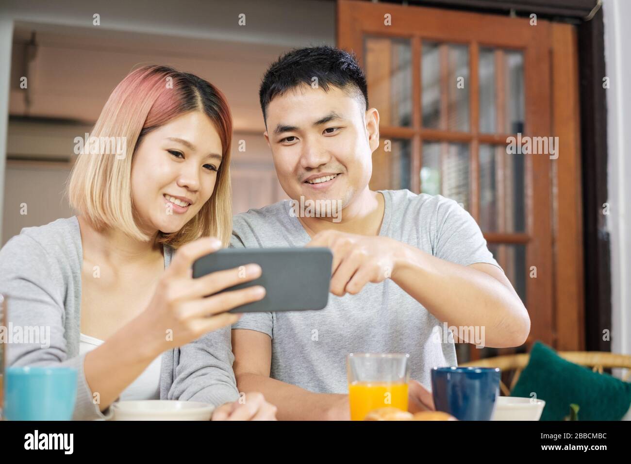 Felice coppia asiatica in pigiama seduti al tavolo in cucina e guardare video sul cellulare togerther a casa la mattina e avente la prima colazione di cereali Foto Stock