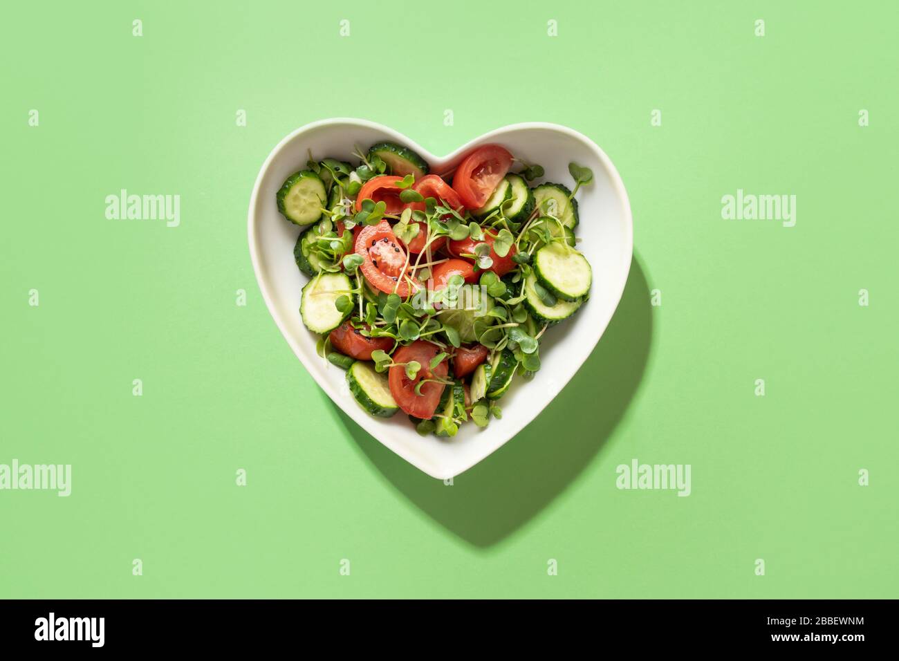 Insalata fresca con pomodoro, cetriolo, verdure, ravanelli microgredi a forma di piatto di cuore sul verde. Vista dall'alto. Concetto vegano e sano mangiare Foto Stock