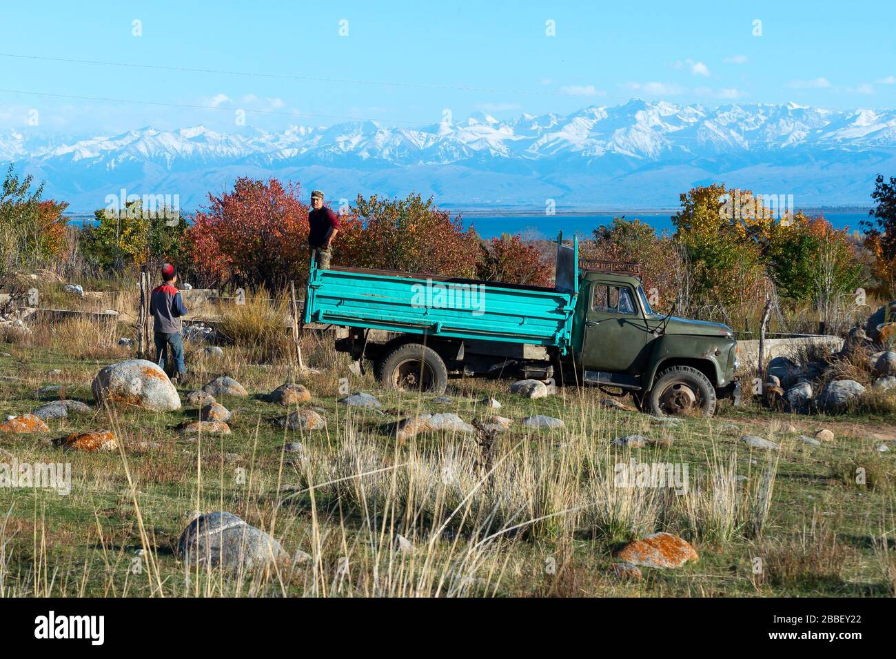 Vecchio camion con due contadini del Kirghizistan. Paesaggio naturale in Kirghizistan con rocce, alberi, Issyk-Kul Lago e montagna con neve. Foto Stock