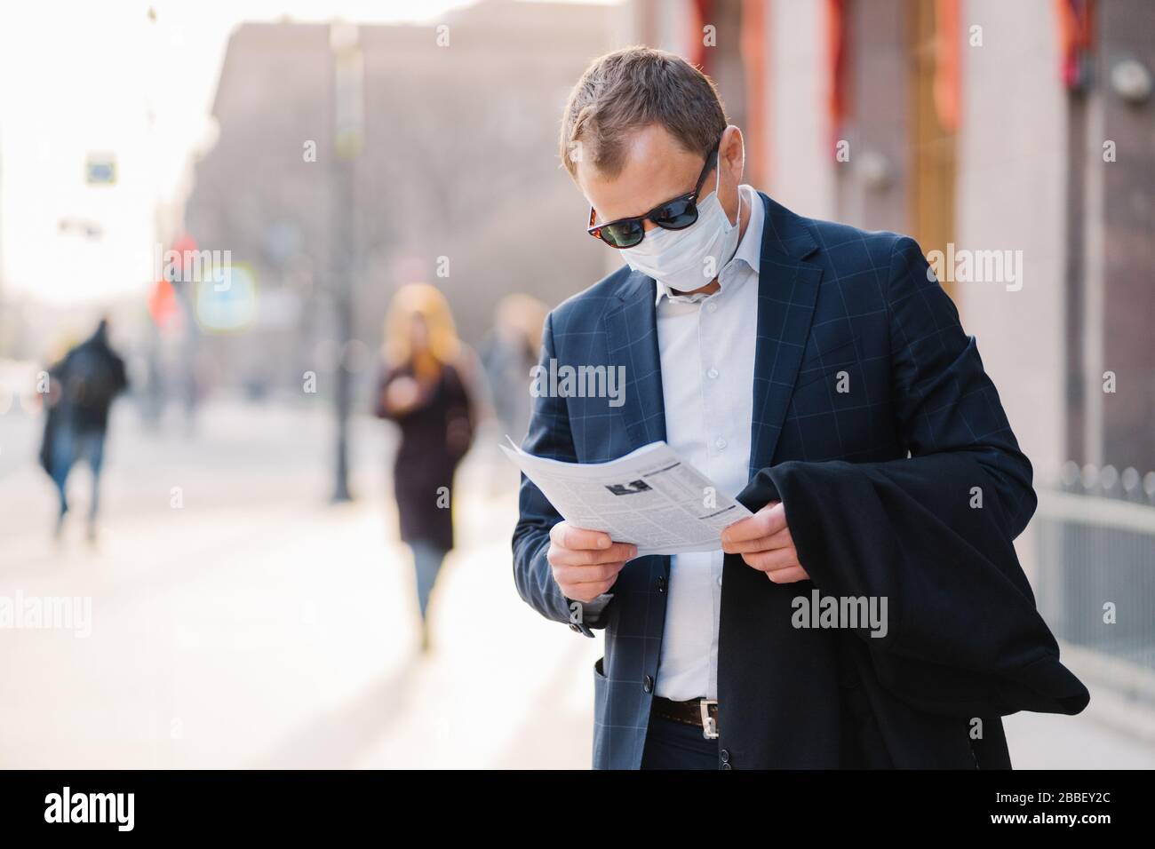 Malattia di coronavirus. Il banchiere maschio serio legge attentamente il giornale, scopre le notizie circa la situazione pandemica intorno al mondo, indossa la maschera protettiva, pre Foto Stock