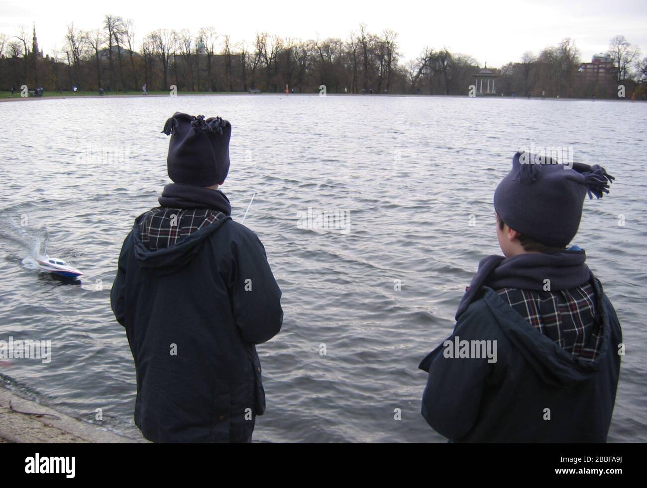 Inverno all'aperto 2 ragazzi con cappotti e sciarpe Motoscafo con telecomando sul lago di Kensington Gardens Inghilterra Regno Unito Foto Stock