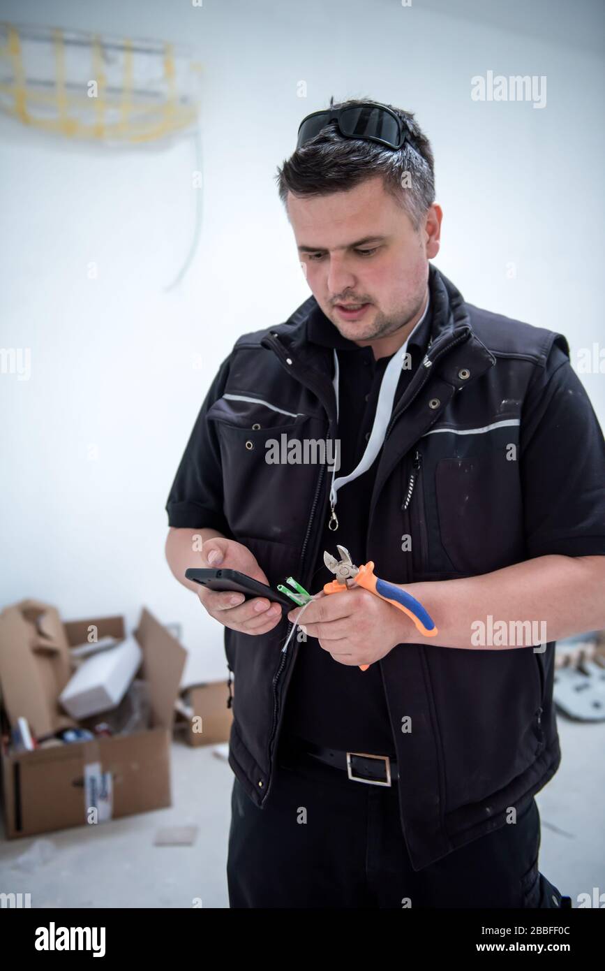 ingegnere elettrico con filo e pinze in mano utilizzando il telefono cellulare mentre si lavora su impianti elettrici in un nuovo appartamento Foto Stock