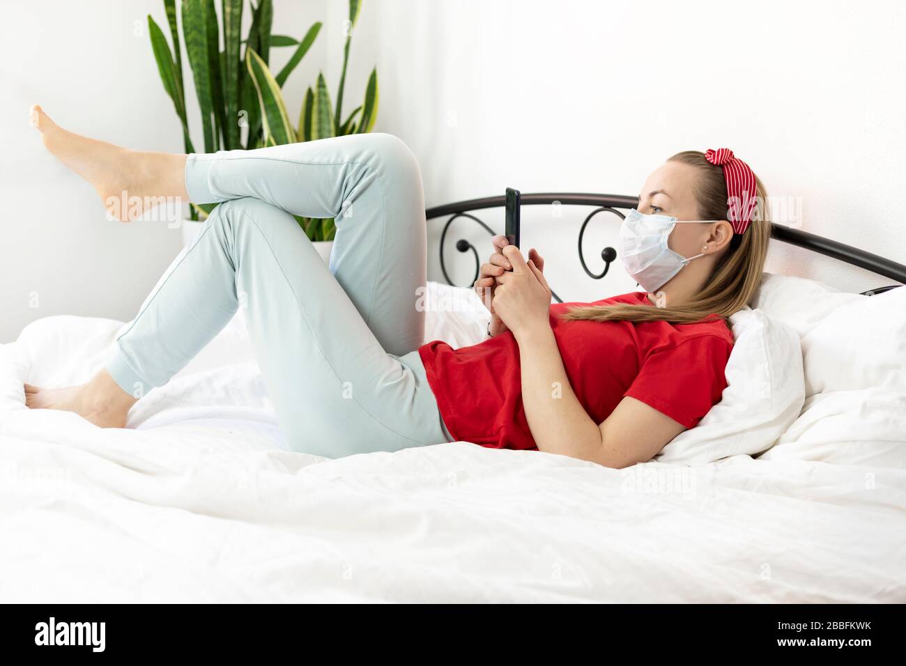 Giovane donna sorridente con capelli biondi in una T-shirt rossa e pantaloni comodi si trova su un letto bianco con uno smartphone. Lavora, studia da casa, scrive un Foto Stock