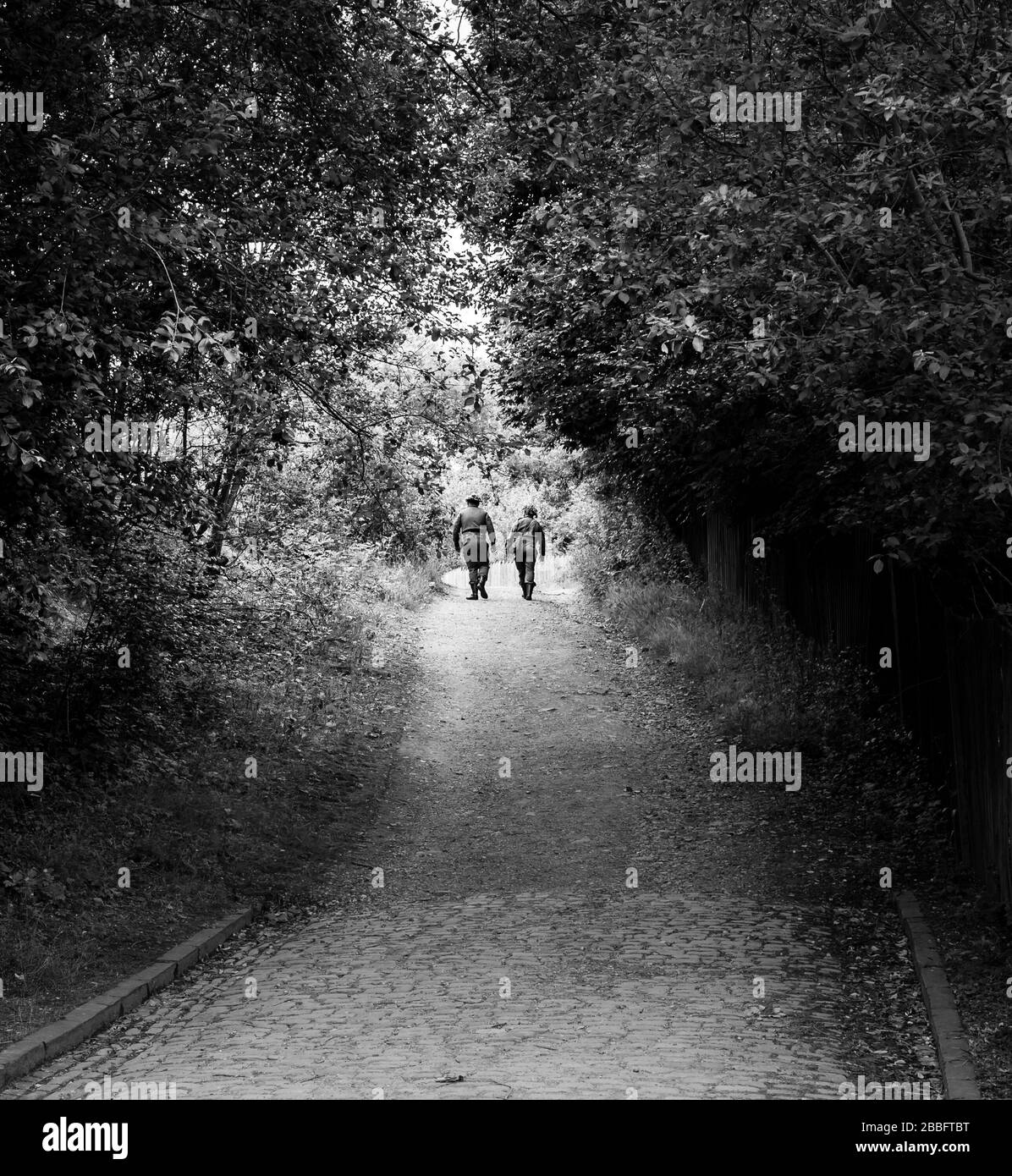 Vista posteriore in bianco e nero di due uomini insieme a distanza in uniforme soldato a piedi lungo la strada tortuosa a 1940s WW2 evento estivo in tempo di guerra, Regno Unito. Foto Stock
