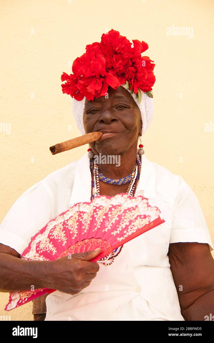 HAVANA, CUBA - 29 MARZO 2017: Ritratto di vecchia donna nera in tipico abbigliamento Havana fumare un grande sigaro cubano su una parete color giallo pastello Foto Stock