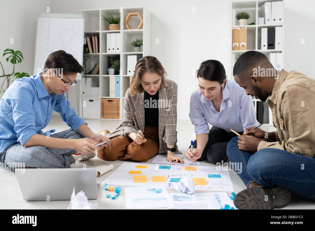 Gruppo di startup interrazziali seduti sul pavimento con task board e l'analisi delle strategie di marketing in ufficio Foto Stock