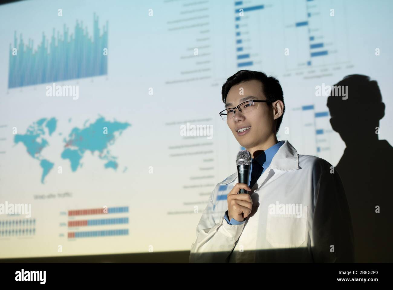 Contenuto giovane biologo asiatico in occhiali e camice da laboratorio che dà voce contro schermo di proiezione con dati statistici Foto Stock