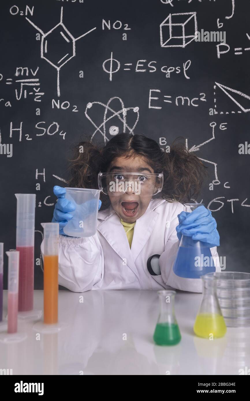ragazza scienziato pazzo con guanti e occhiali in camice da laboratorio con palloni chimici, sfondo lavagna con formule scientifiche, esplosione nel laboratorio Foto Stock