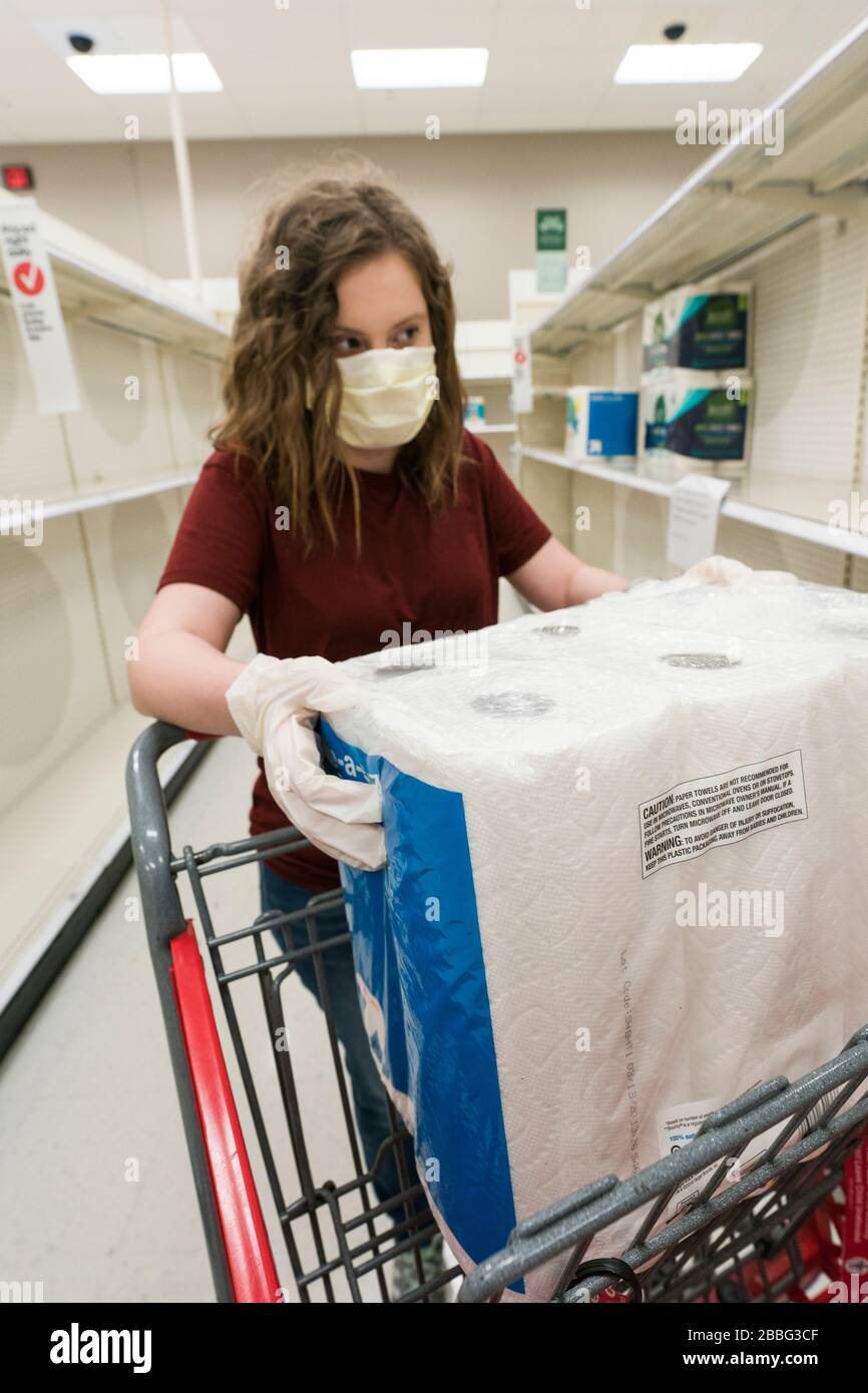 Indiana, USA - 24 marzo 2020: Una donna bionda che indossa una maschera facciale e guanti mette un pacchetto di asciugamani di carta nel suo carrello in mezzo a scaffali vuoti Foto Stock