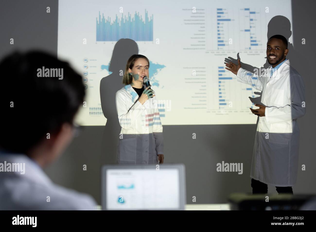 Intraprendenti tirocini multietnici in camici di laboratorio che presentano informazioni mediche utilizzando lo schermo di proiezione alla conferenza scientifica Foto Stock