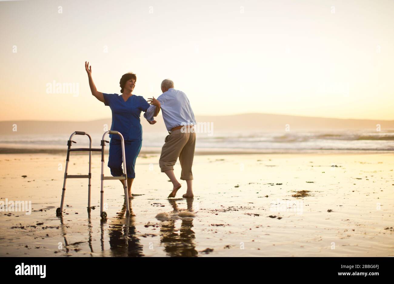 L'uomo anziano e un'infermiera femminile ballano insieme accanto ad un aiuto per camminare sulla spiaggia. Foto Stock