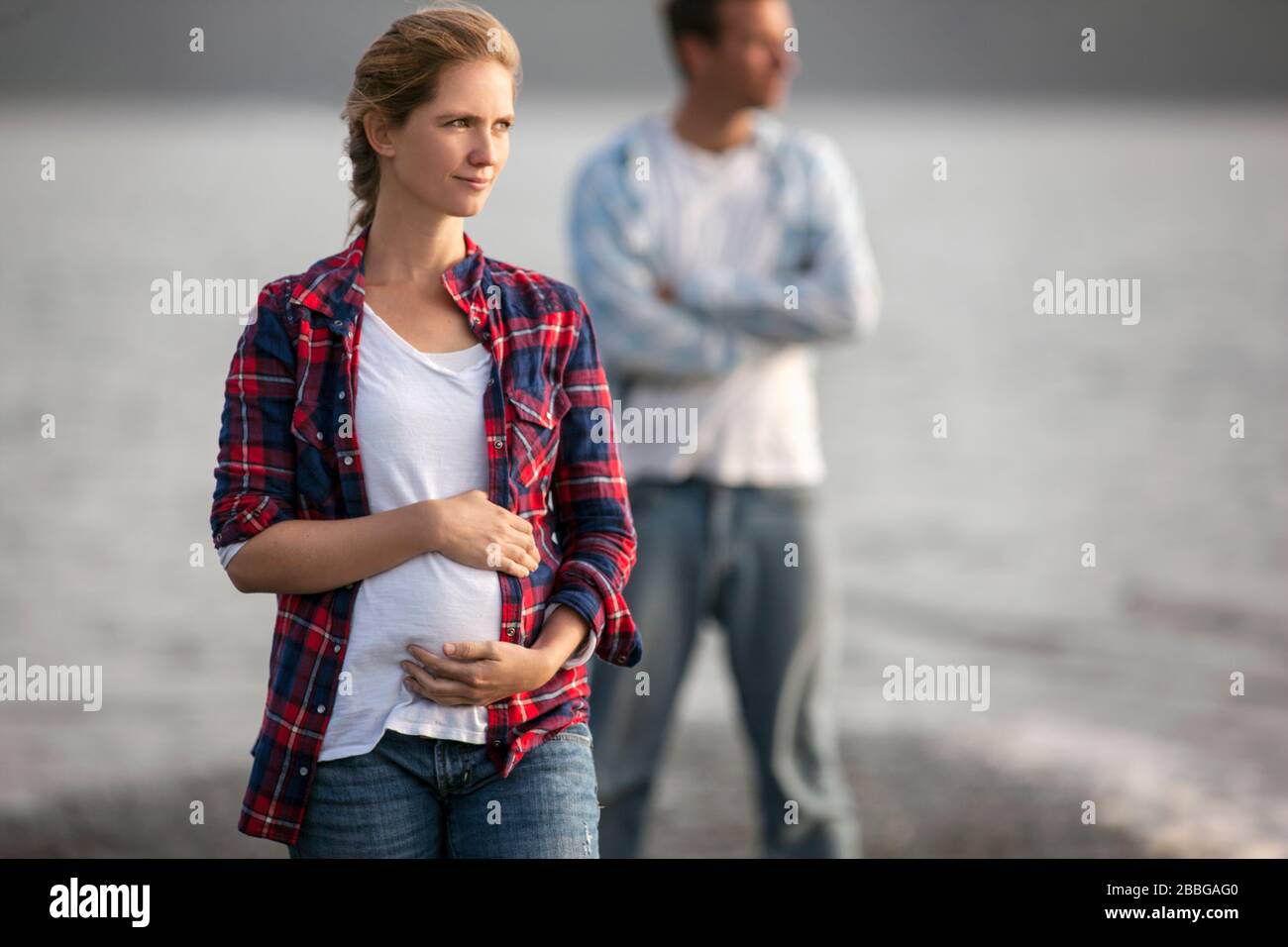 Ritratto di una donna incinta in spiaggia tenendola delicatamente il ventre con il marito in piedi sullo sfondo Foto Stock