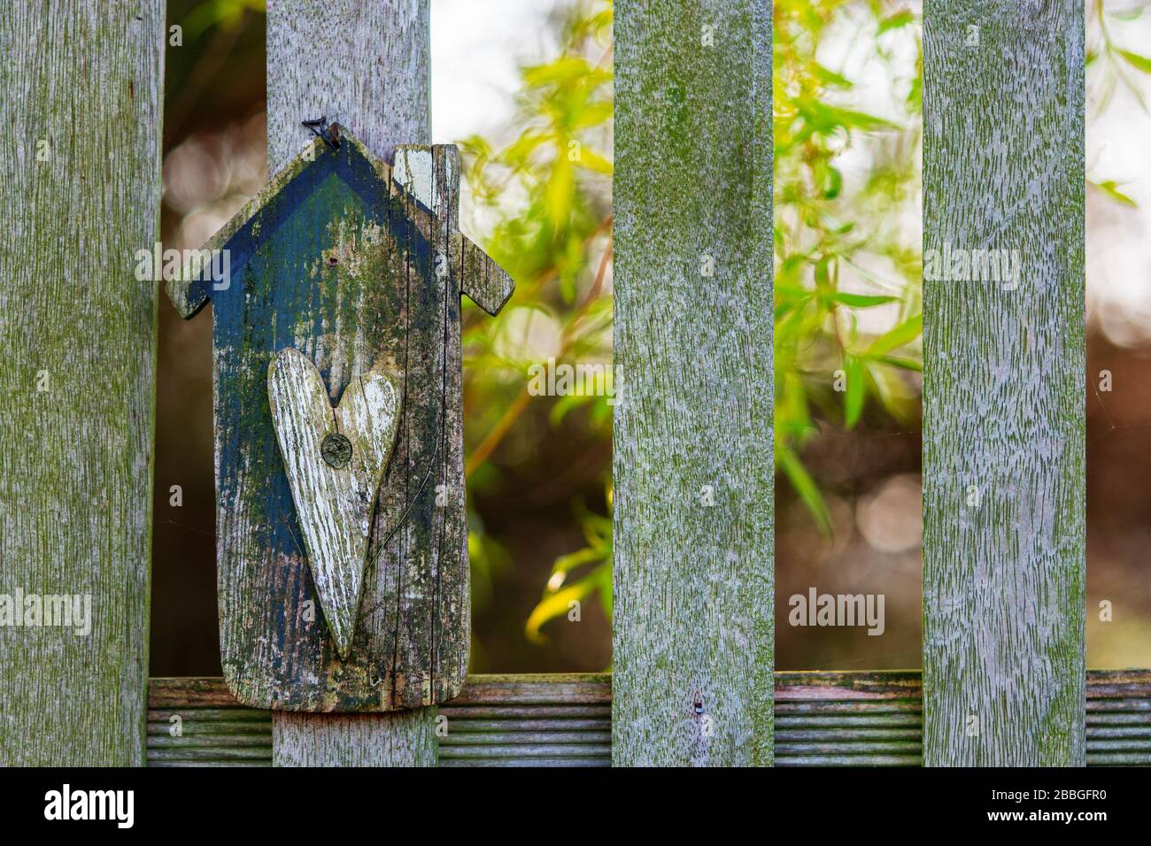 Pannello di legno a forma di casa lavorato a mano con forma a cuore su una recinzione da giardino. Albero con foglie verde brillante su sfondo soleggiato. Casa, concetto giardino Foto Stock