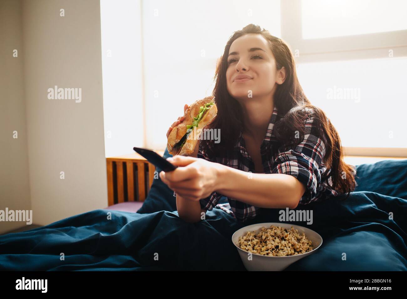 Giovane bella donna nel letto del mattino a casa. Una donna entusiasta che guarda la tv. Utilizzando il telecomando e mangiando sandwich con popcorn. Film Foto Stock