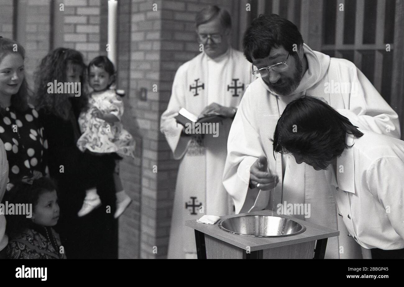 1970s, storico, all'interno di una chiesa, i membri della famiglia che guardano una donna che si fa battezzare, come un sacerdote robed ama la sua testa, Londra, Inghilterra, Regno Unito. Il battesimo è un rito cristiano di ammissione, un sacramento, un'ordinanza di Gesù Cristo. Foto Stock