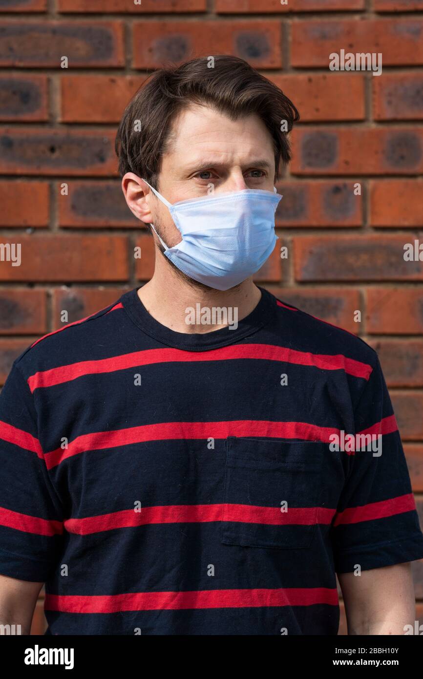 Metà 40's maschio casual indossa maschera viso blu a altezza di Coronavirus focolaio, contro muro di mattoni. Foto Stock