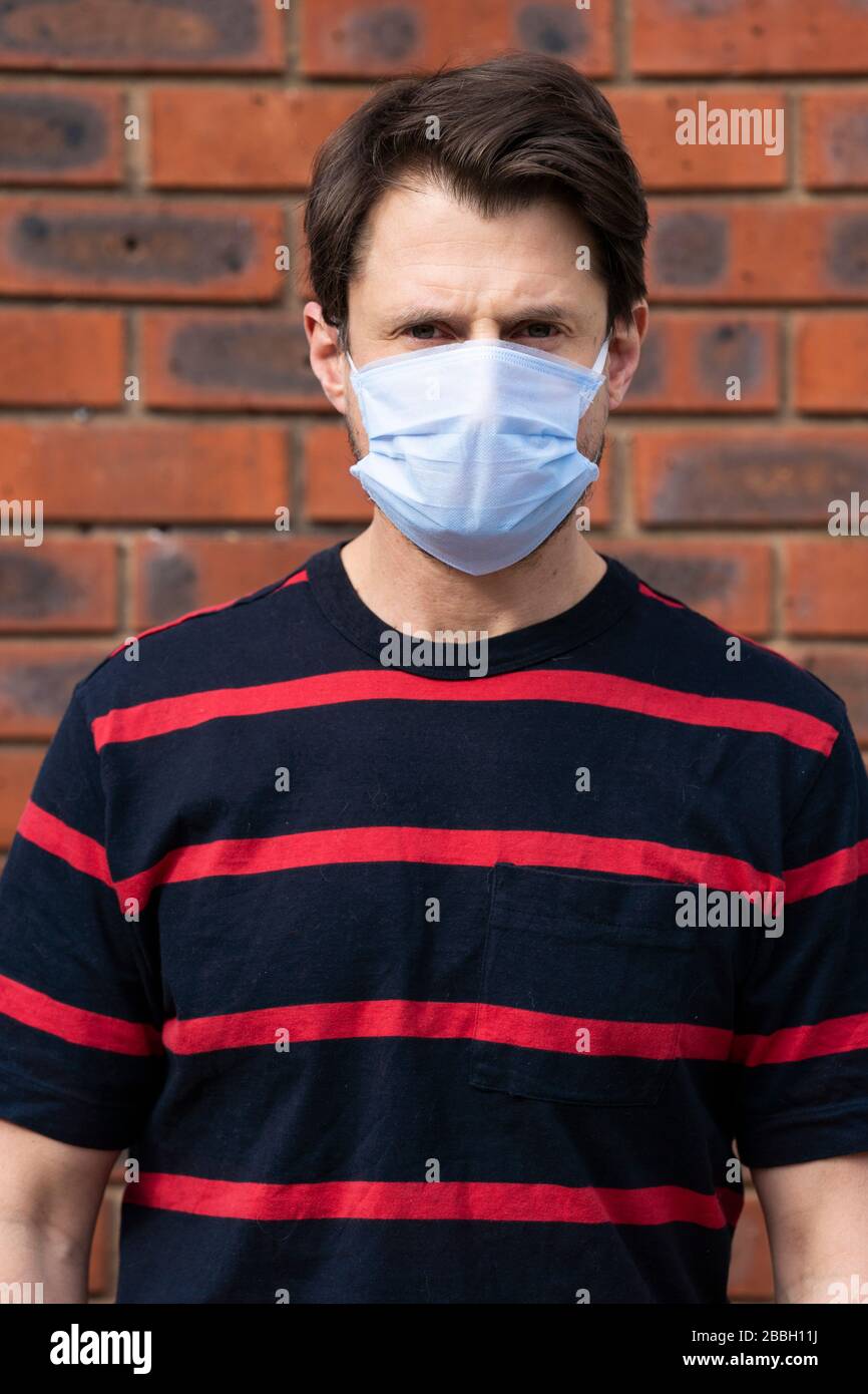 Metà 40's maschio casual indossa maschera viso blu a altezza di Coronavirus focolaio, contro muro di mattoni. Foto Stock