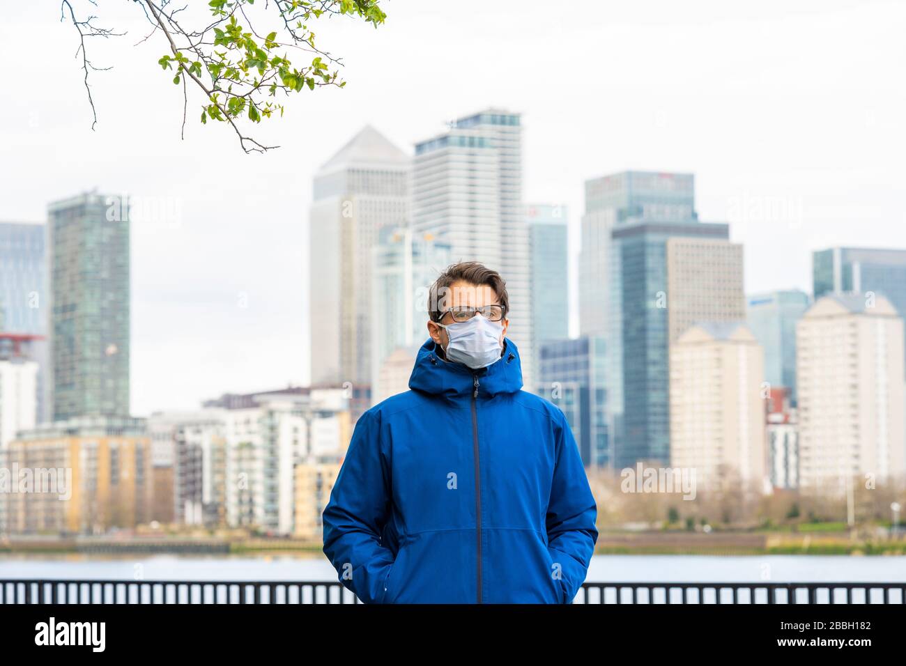 A metà degli anni '40 uomini indossano giacca blu e maschera facciale passeggiate lungo il Tamigi, con il quartiere finanziario di Londons e Canary Wharf sullo sfondo. Foto Stock