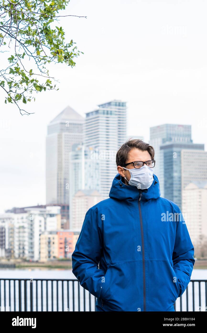 A metà degli anni '40 uomini indossano giacca blu e maschera facciale passeggiate lungo il Tamigi, con il quartiere finanziario di Londons e Canary Wharf sullo sfondo. Foto Stock