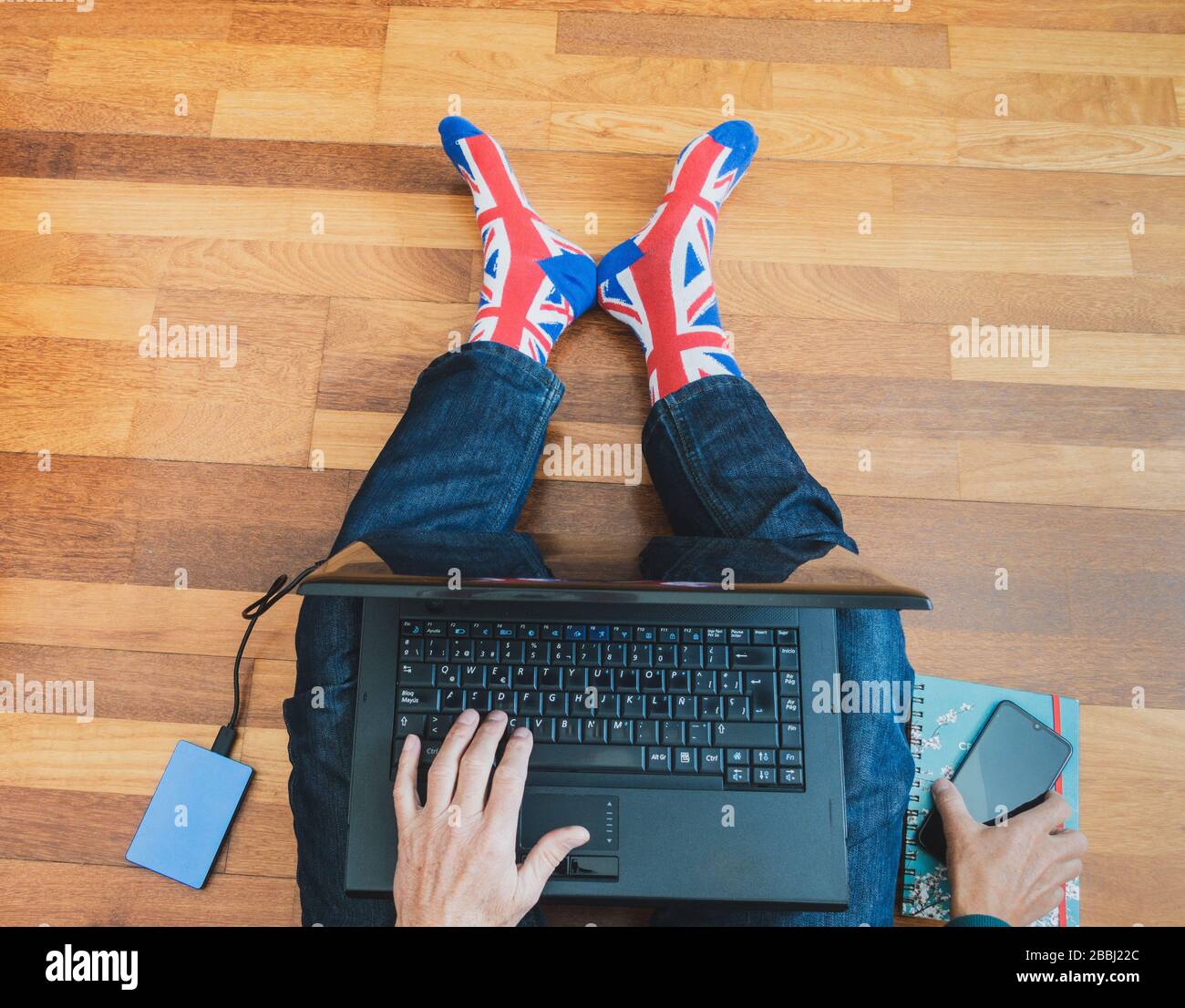 Uomo che indossa una calza jack a bandiera a Unione seduta su pavimento in legno con laptop. Lavorare da casa, auto isolamento, allontanamento sociale, Coronavirus... concetto Foto Stock
