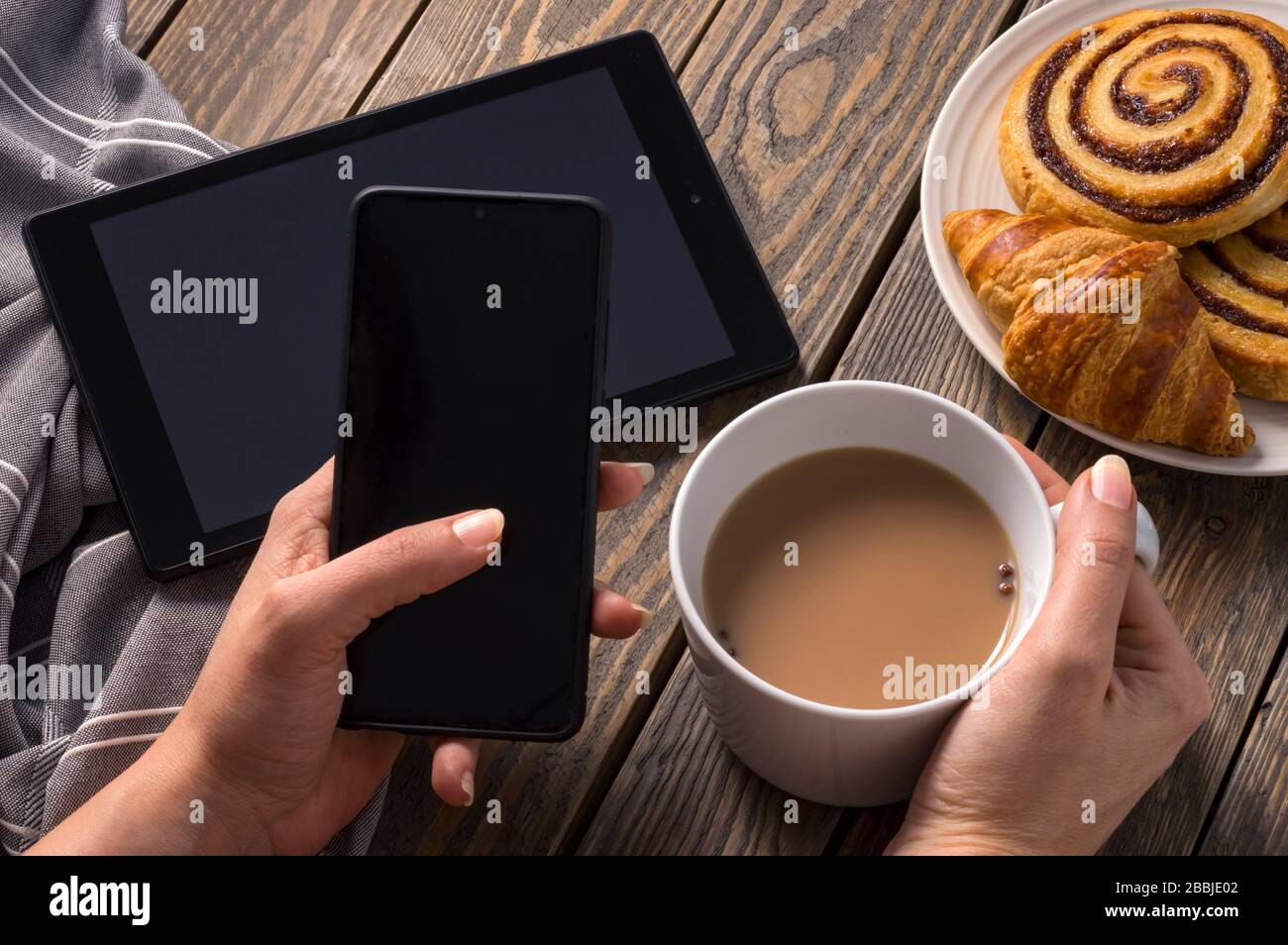 Una donna che fa colazione su un tavolo rustico che controlla le ultime novità sui suoi dispositivi elettronici mentre ha tè e pasticceria. Scorrere con il pollice sinistro acceso Foto Stock