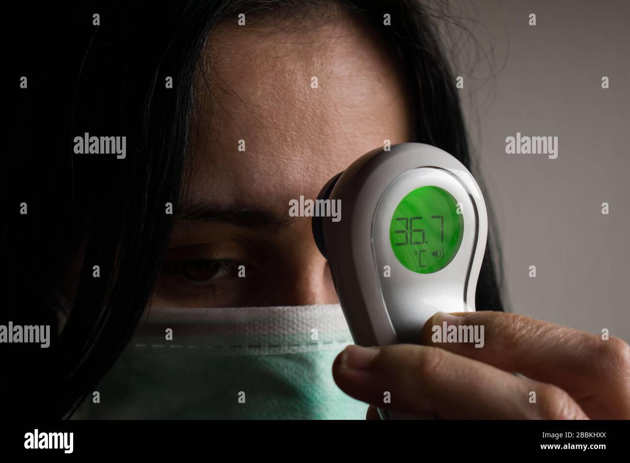 La giovane donna preoccupata che indossa una maschera protettiva per il viso medica misura la temperatura con un termometro elettronico. Concetto di quarantena dei virus o dell'influenza Foto Stock