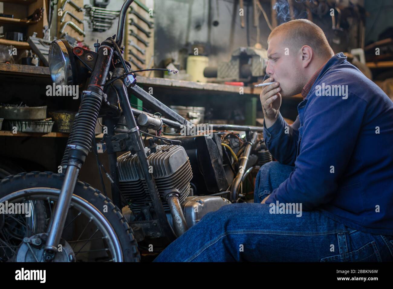 L'uomo fuma durante una pausa di riparazione della motocicletta in officina Foto Stock