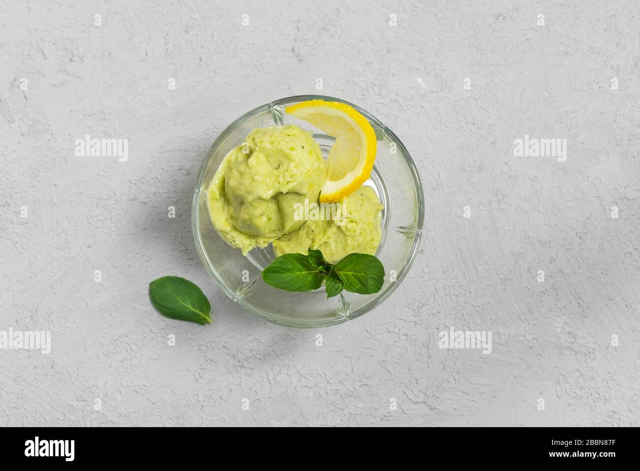 Dolce vegano fatto in casa gelato avocado con foglie di limone e menta in una ciotola di vetro su sfondo grigio. Vista dall'alto con spazio di copia Foto Stock
