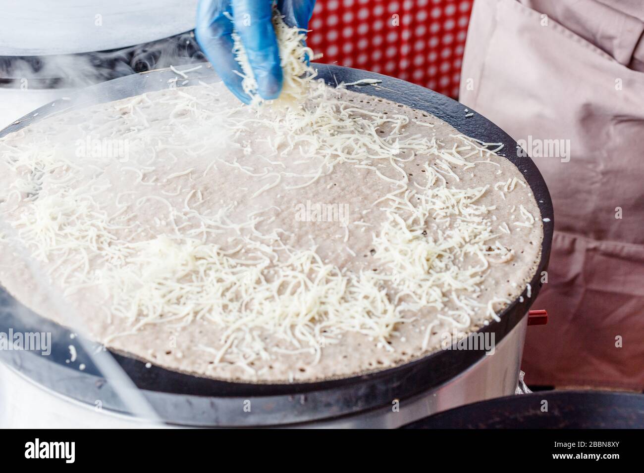 Fare crepes frittelle in un mercato alimentare. Una mano sta facendo crepes saporite all'aperto aggiungendo il formaggio grattugiato alla crepe su un grlicdle di metallo. Foto Stock