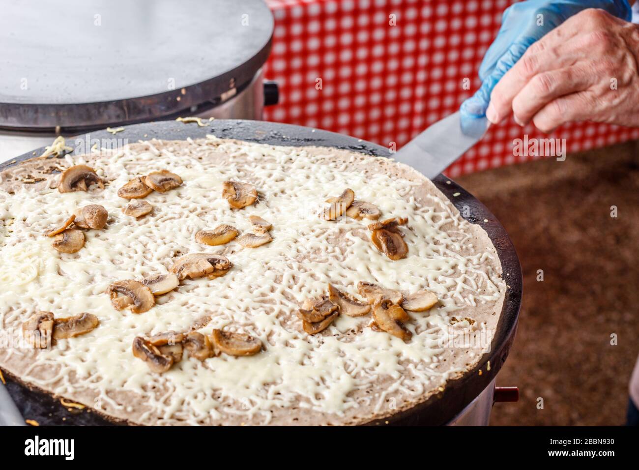 Fare crepes frittelle in un mercato alimentare. Una mano sta facendo crepes saporite all'aperto con il formaggio fuso e i funghi su una griglia metallica. Foto Stock