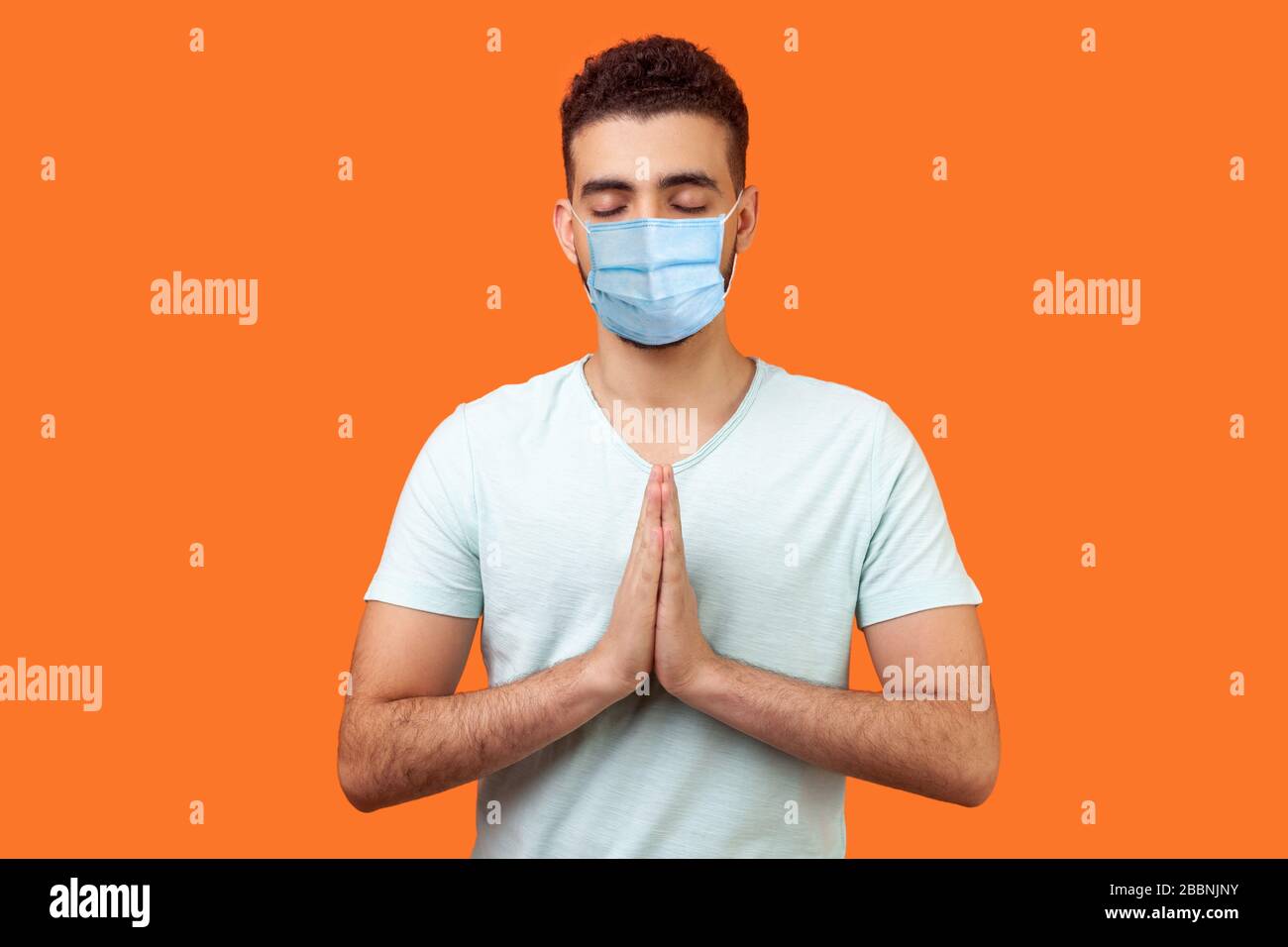 Pratica yoga per equilibrio mentale. Ritratto di uomo pacifico con maschera medica chirurgica in t-shirt bianca che tiene le mani in namaste o preghiera, meditando Foto Stock