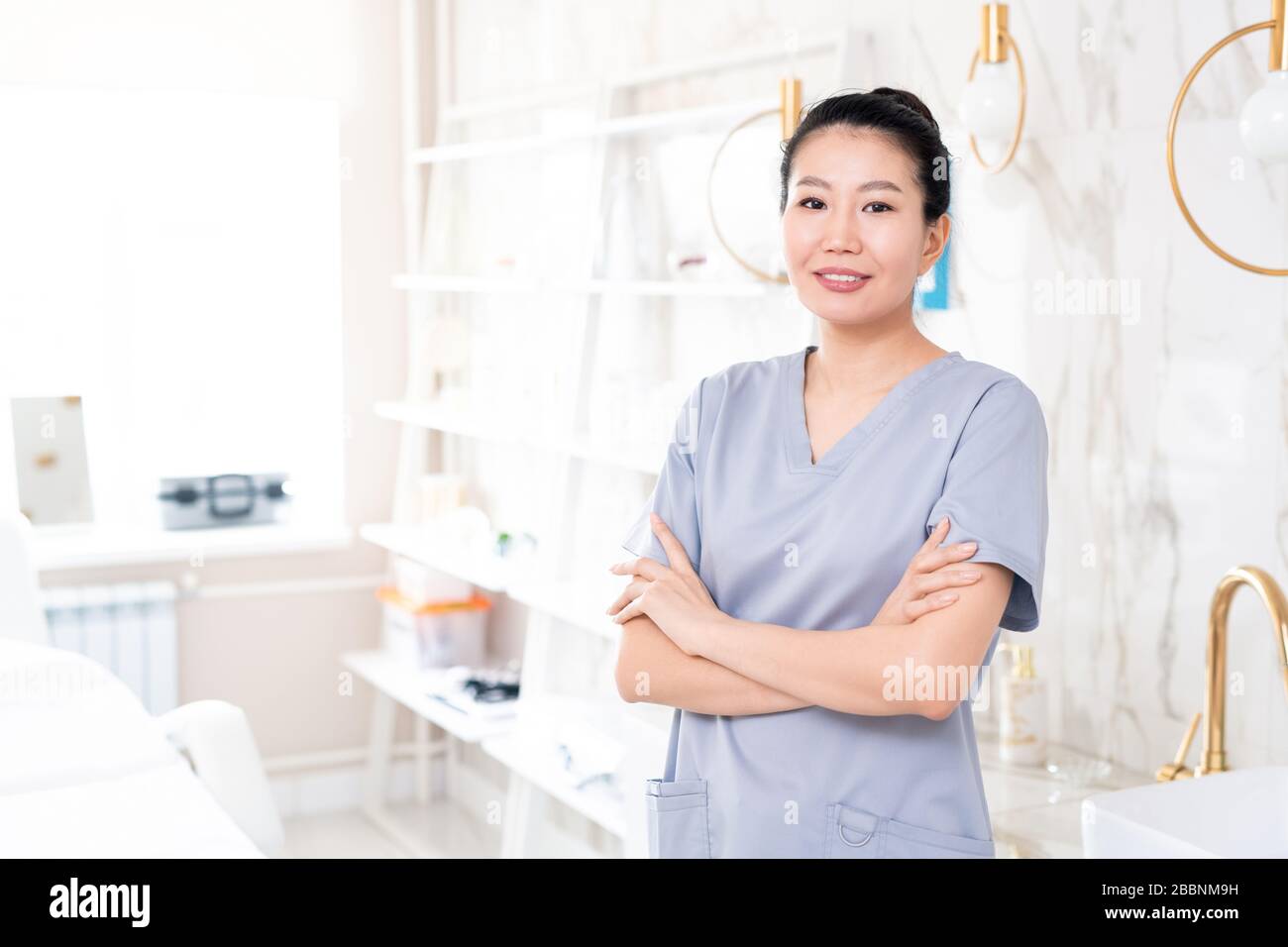 Ritratto di estetista asiatico positivo di mezza età in scrub in piedi con braccia incrociate in sala trattamento di bellezza con design contemporaneo Foto Stock