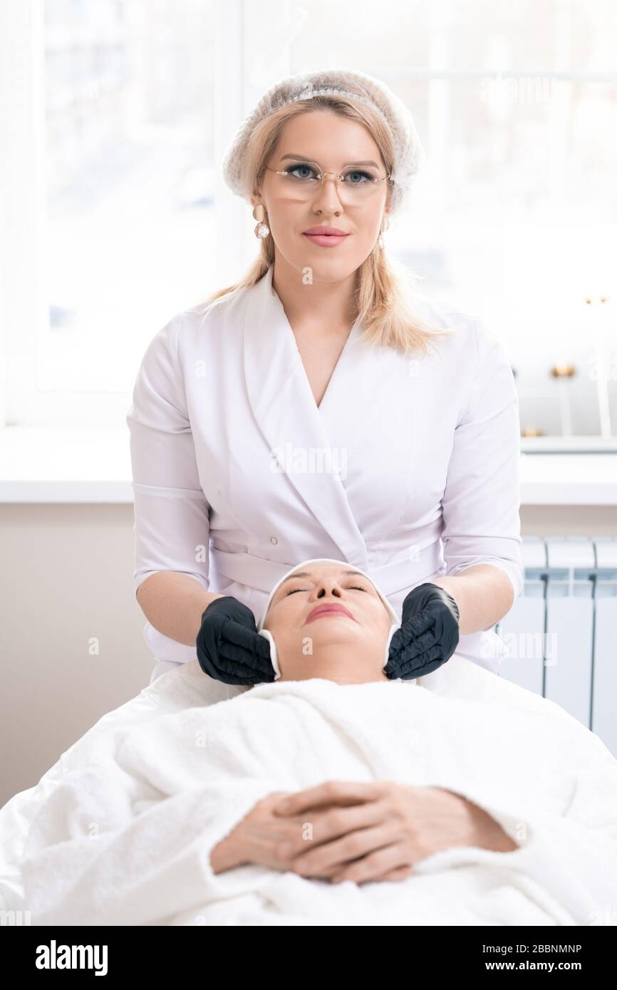 Ritratto di colometologo giovane in guanti neri con batuffolo di cotone mentre si pulisce la pelle del viso di donna matura prima della procedura Foto Stock