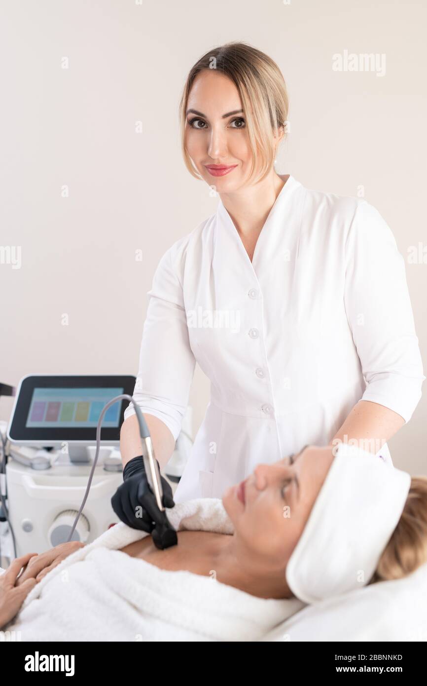 Ritratto di contenuto sicuro specialista di bellezza in camice da laboratorio utilizzando la macchina cosmetologica per lisciare la pelle sul petto dei clienti Foto Stock