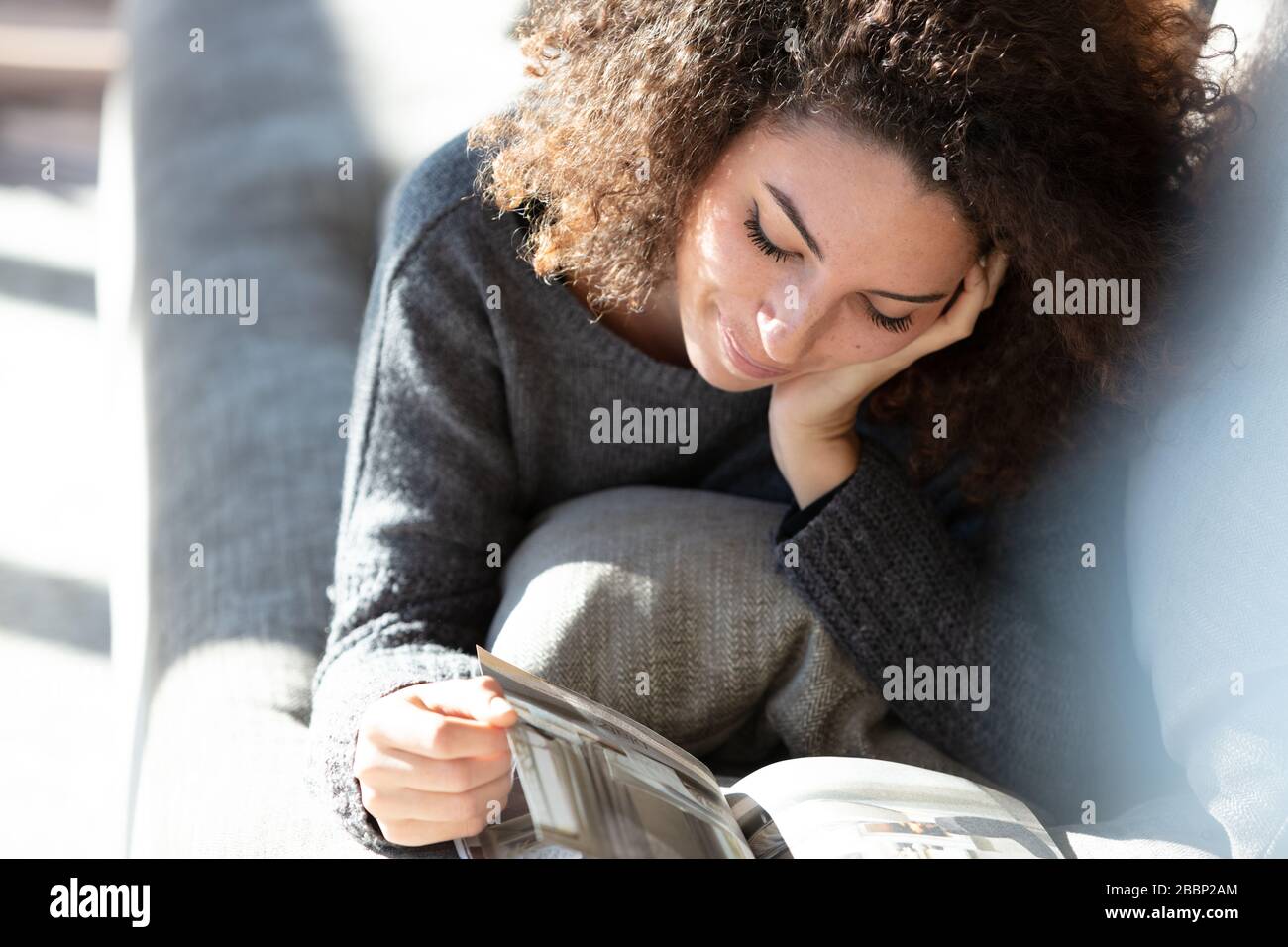 Una donna piuttosto giovane sdraiata su un moderno salotto leggendo una rivista al sole del pomeriggio. Foto Stock