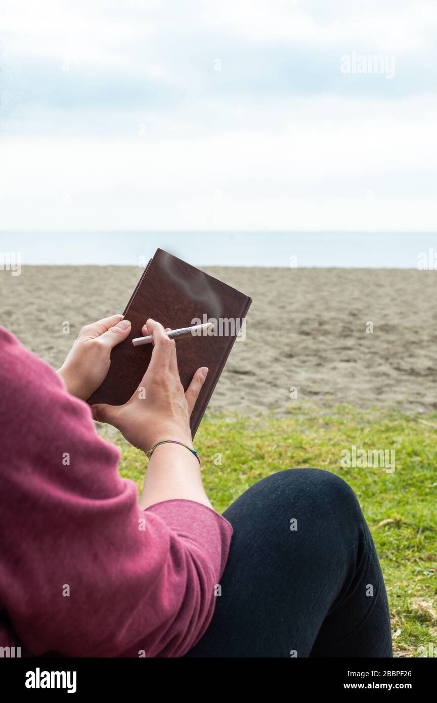La persona che legge un libro sulla spiaggia prende una pausa per fumare un giunto o una sigaretta di marijuana. Sfocatura sfondo con mare e cielo. Foto Stock