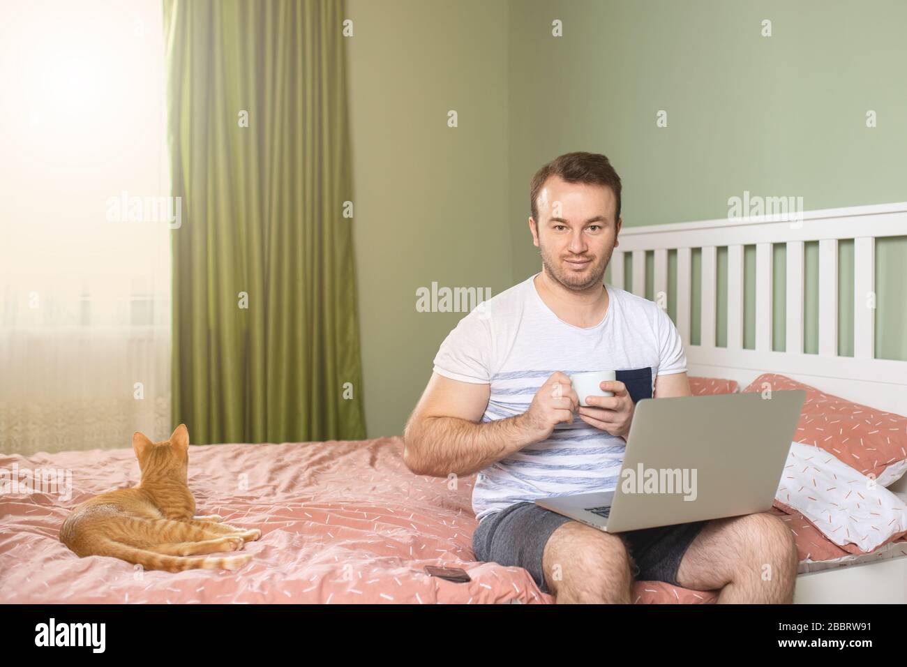 Giovane con un computer portatile a letto che lavora a casa con una tazza di caffè. Un allegro adulto che lavora dal suo letto di appartamento con un gatto accanto a lui sul letto. Foto Stock