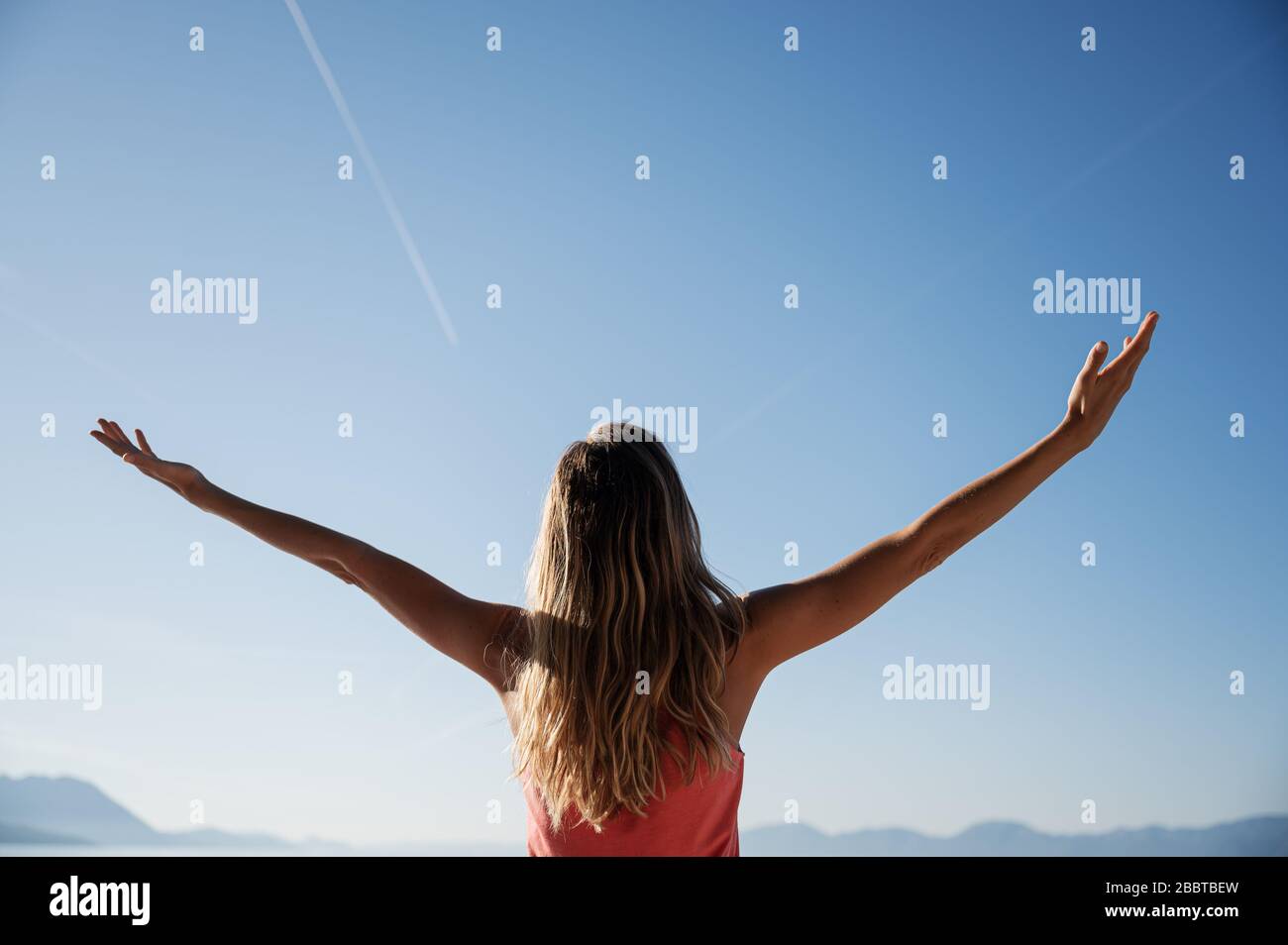 Vista da dietro di una giovane donna con lunghi capelli in piedi fuori sotto cielo estivo chiaro con le braccia sollevate e diffusione. Foto Stock