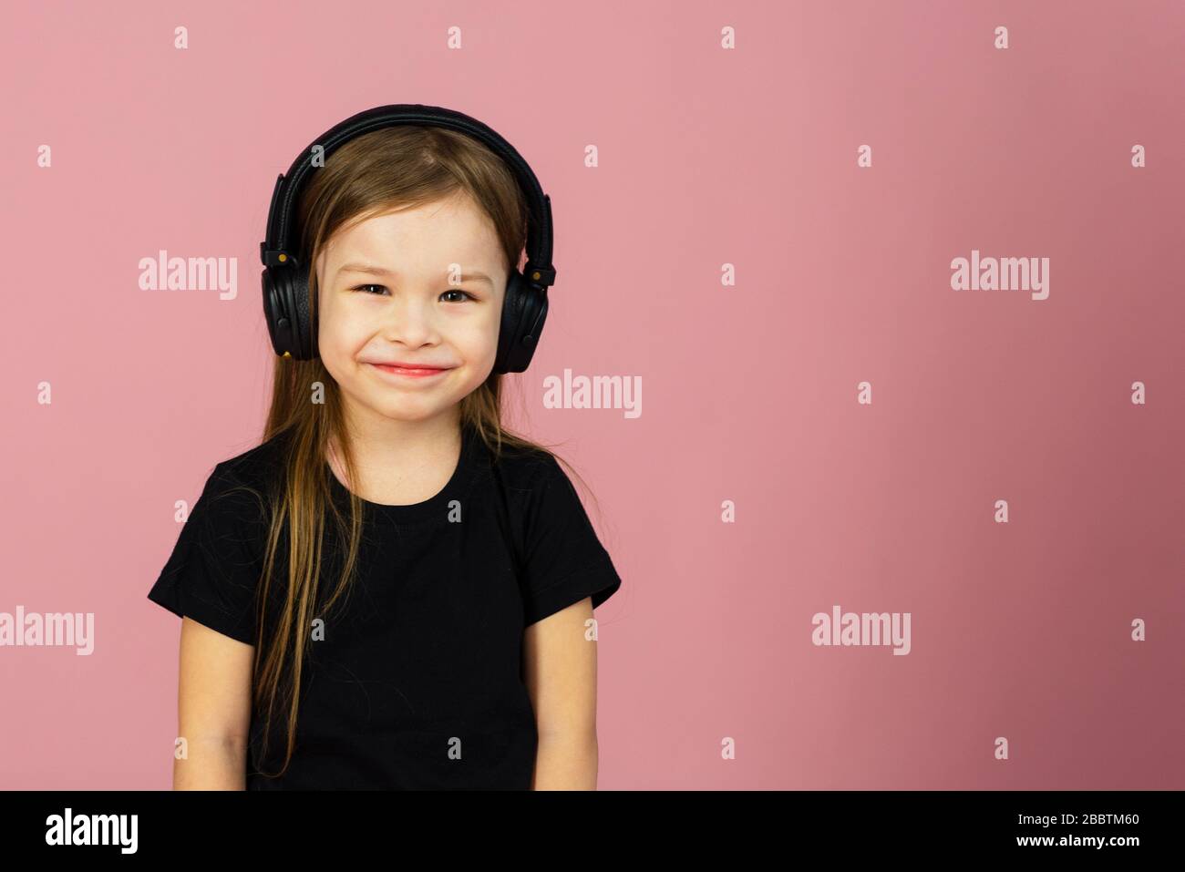 Una bambina in wireless grandi cuffie nere su uno sfondo rosa pastello guarda la fotocamera. Il concetto di ascolto e di ascolto della musica. Luogo per il testo, copyspace. Foto Stock