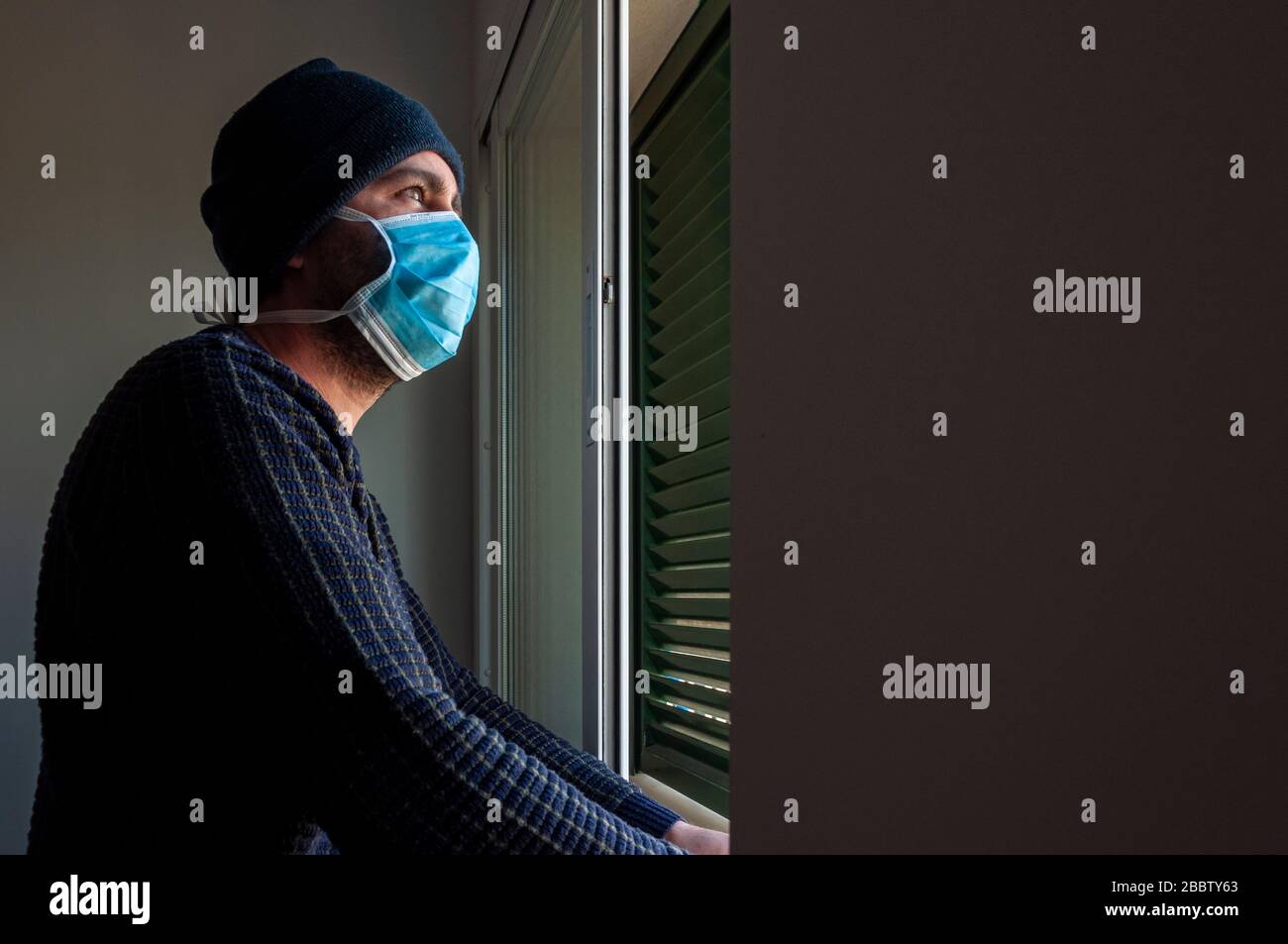 Ragazzo con maschera blu guarda da un interno scuro nella luce della finestra. Bloccato nella casa da confinamento. Coronavirus. Covid-19 Foto Stock
