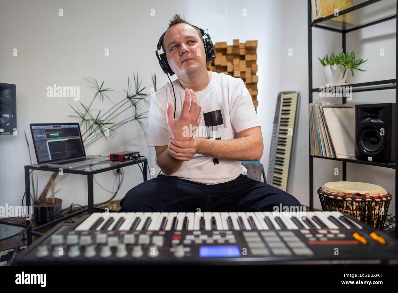 Musicista professionista che ha dolore al polso durante la riproduzione della tastiera midi in studio digitale a casa. È circondato da strumenti e controller midi. Foto Stock