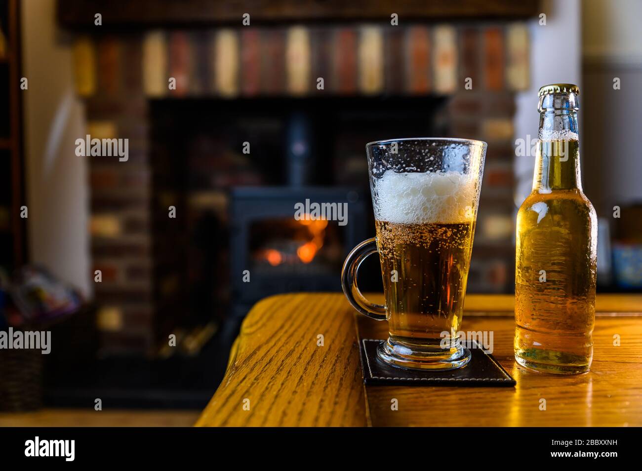 Birra versata in vetro su tavola di quercia con fuoco di legna su sfondo a casa Foto Stock